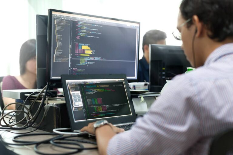 Photo of man at a laptop and computer desk, writing lines of code