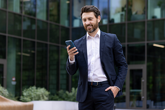 A smiling young man in a business suit is walking near an office building rubbing his hand in his pocket and using a mobile phone